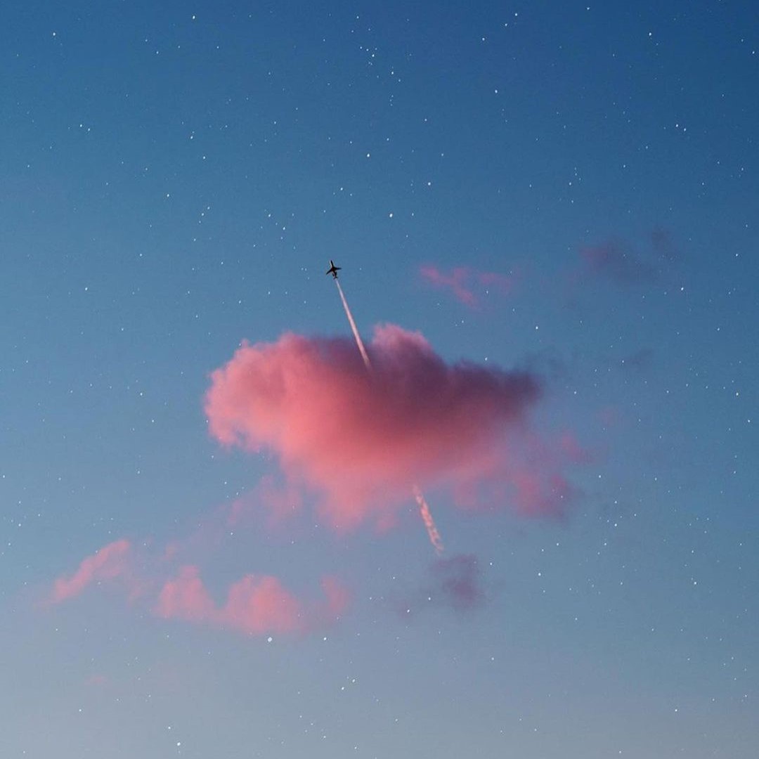 A single pink cloud in a blue evening sky, pierced by a jet and its contrail.