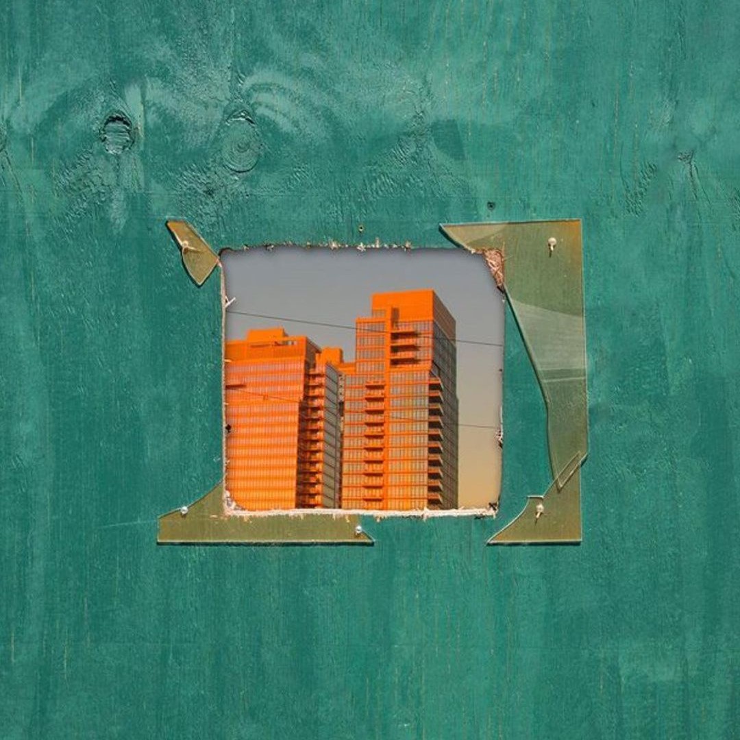 Orange apartment towers seen through a hole cut in a green plywood hoarding.