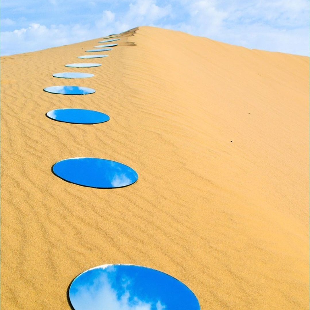 Round mirrors laid in a line up a sand dune, each one holding a piece of the sky.