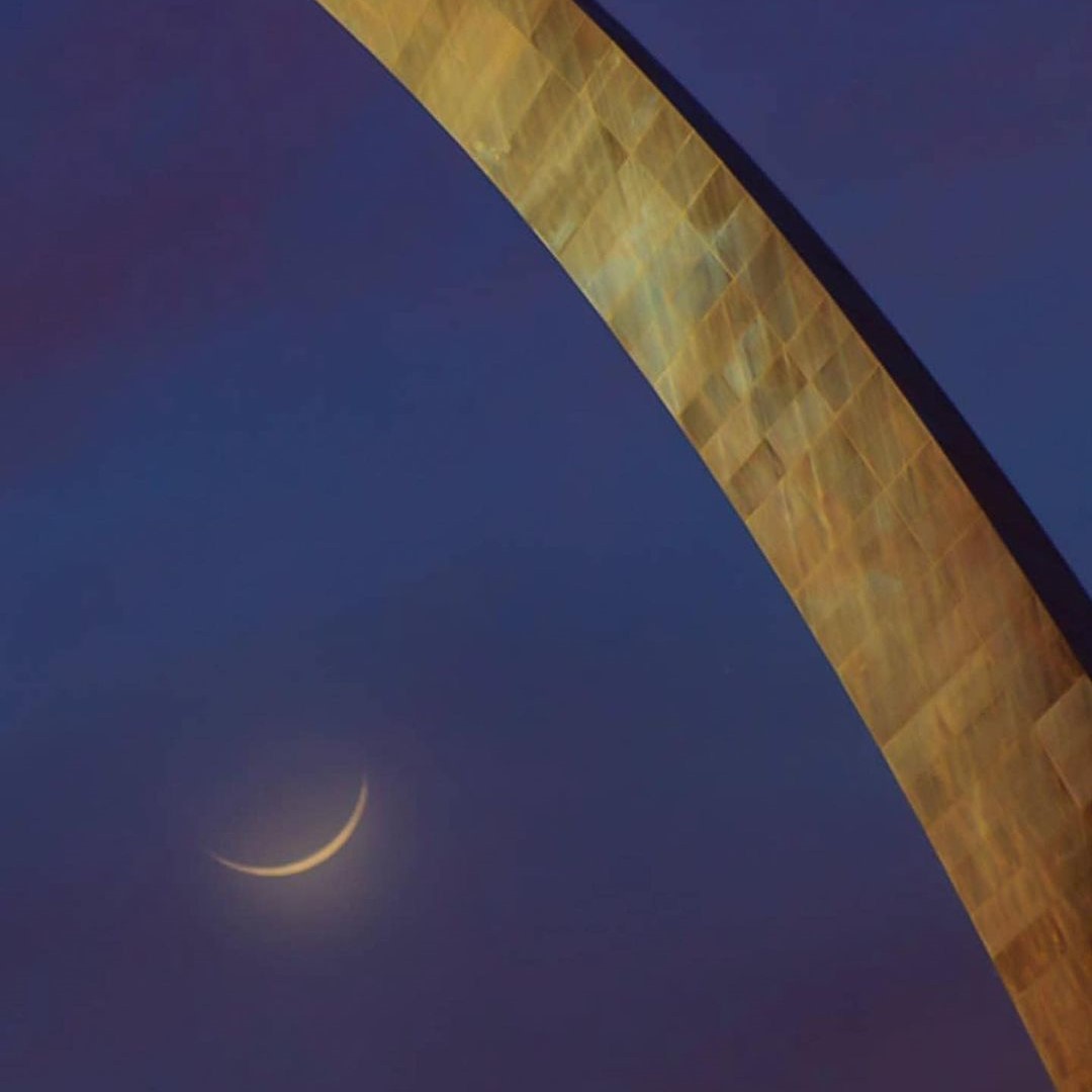 The Gateway Arch cutting across a deep blue sky beside a crescent moon.