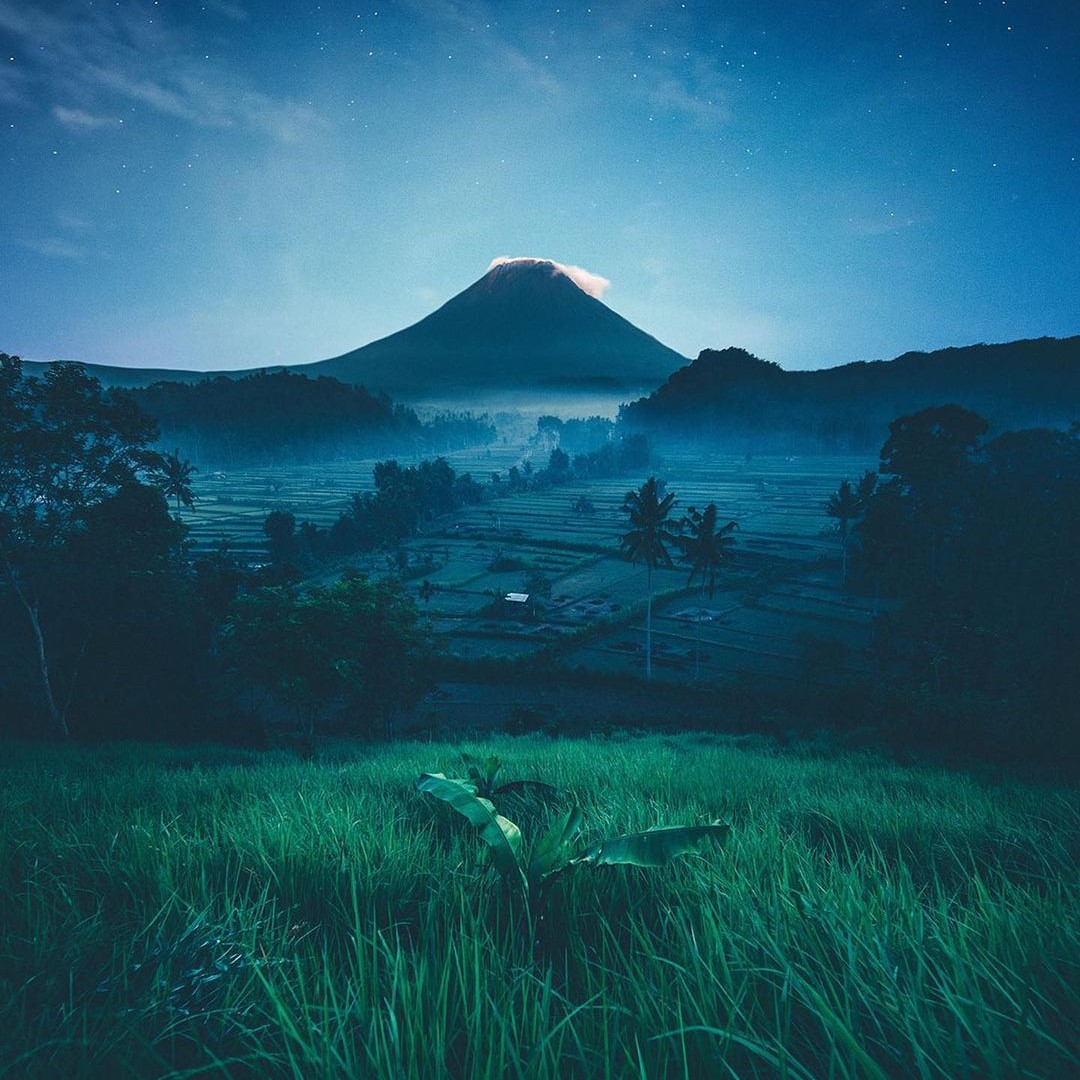 A volcano above terraced fields at night, everything lit blue.